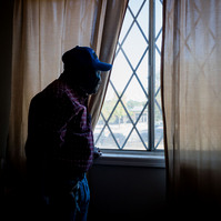 A man wears a plaid shirt and ball cap as he looks out of a window.