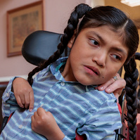 A boy in a striped shirt sits in a wheelchair with his hands in front of him.