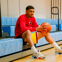 A young African American man sits on bleachers in a gym holding a basketball. 