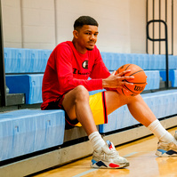 A young African American man sits on bleachers in a gym holding a basketball. 