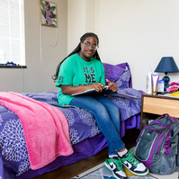 Young African American woman wears braids and a green shirt as she sits on a colorful bed in her dorm room. 