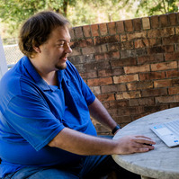 A man in a blue polo sits at a table and works on a laptop computer.