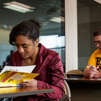African American woman wears a rose jacket as she works in a yellow text book during class. 