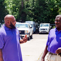 Two African American men wear matching purple polo shirts as they stand and talk in a parking lot. 