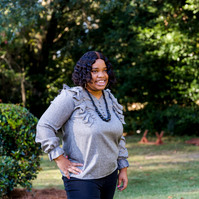 A young woman with dark hair wears a grey blouse and a black necklace. She is standing outside in front of trees and is smiling. 