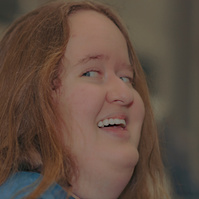 Woman with red hair wears a blue t-shirt and smiles at the camera over her shoulder. 