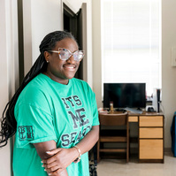 Young African American woman wears braids and a green shirt while standing in a dormitory hallway.