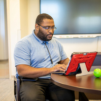 A young black male wears a blue shirt and glasses as he sits at a table and works on a computer. 