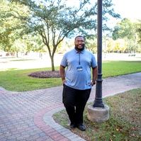 A young black male wears glasses and a blue shirt and stands on a tree-lined path.