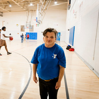 A woman with brown hair stands inside a gymnasium with basketball players on the court. 