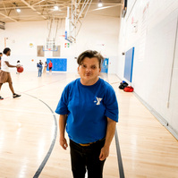 A woman with brown hair stands inside a gymnasium with basketball players on the court. 