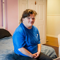 A woman with brown hair wears a blue shirt and sits on the end of a bed.