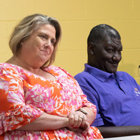 A man and a woman wear bright shirts and sit at a table in front of a yellow wall. 