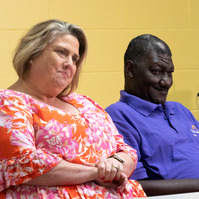 A man and a woman wear bright shirts and sit at a table in front of a yellow wall. 