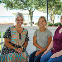 Three women sit on a bench outside and smile for the camera.