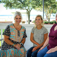 Three women sit on a bench outside and smile for the camera.