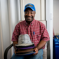 African American man in a plaid shirt and blue ball cap smiles in his living room while holding a stack of hats.