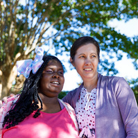 Mother and daughter stand outside under a tree on a sunny day. 