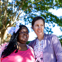 Mother and daughter stand outside under a tree on a sunny day. 