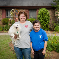 Two women stand in front of a house as one of them holds a dog. 