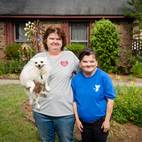 Two women stand in front of a house as one of them holds a dog. 