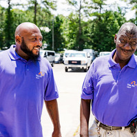 Two African American men wear matching purple polo shirts as they stand and talk in a parking lot. 