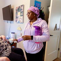 A young brunette woman in a patterned shirt and glasses takes medication from a care attendant wearing a pink jacket and headwrap.