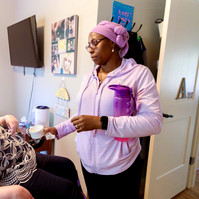 A young brunette woman in a patterned shirt and glasses takes medication from a care attendant wearing a pink jacket and headwrap.
