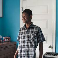 A young African American man in a plaid shirt stands in his blue bedroom. 
