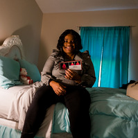 A young woman in a grey blouse sits on a pastel-colored bed in a bedroom.