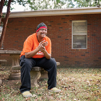 A man in an orange shirt and knit cap sits outside on a bench smiling.