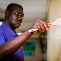 A middle aged African American man wears a purple polo shirt and paints a wall cream.