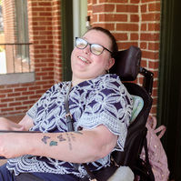 A young brunette woman in a patterned shirt and glasses holds a book and sits on a screened porch. 