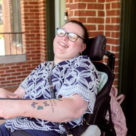 A young brunette woman in a patterned shirt and glasses holds a book and sits on a screened porch. 