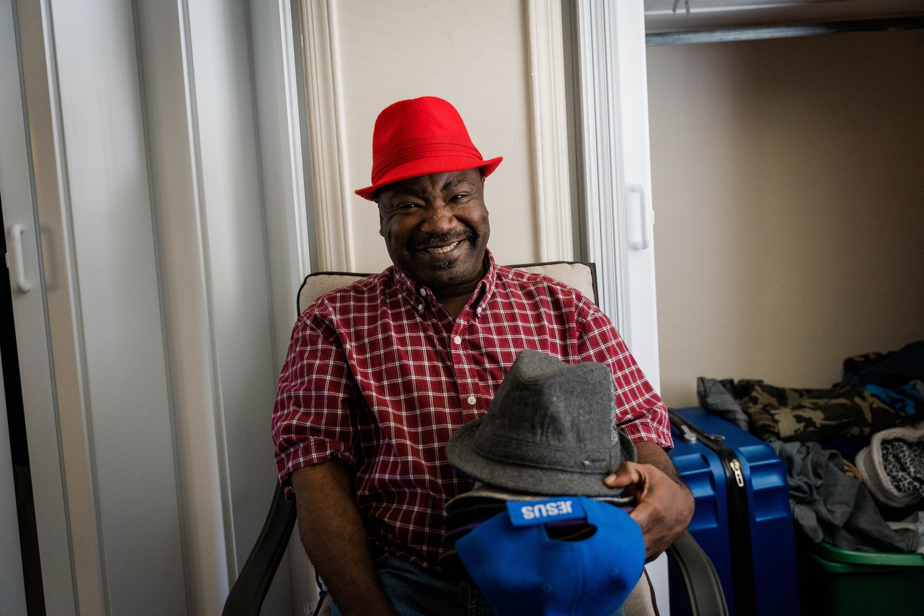 African American man in a plaid shirt and red hat smiles in his living room while holding a stack of hats.
