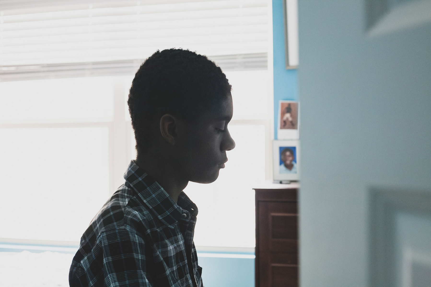 A young African American man in a plaid shirt sits in front of a window in his room. 