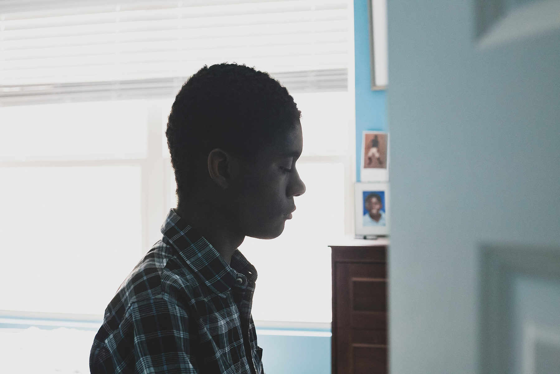 A young African American man in a plaid shirt sits in front of a window in his room. 