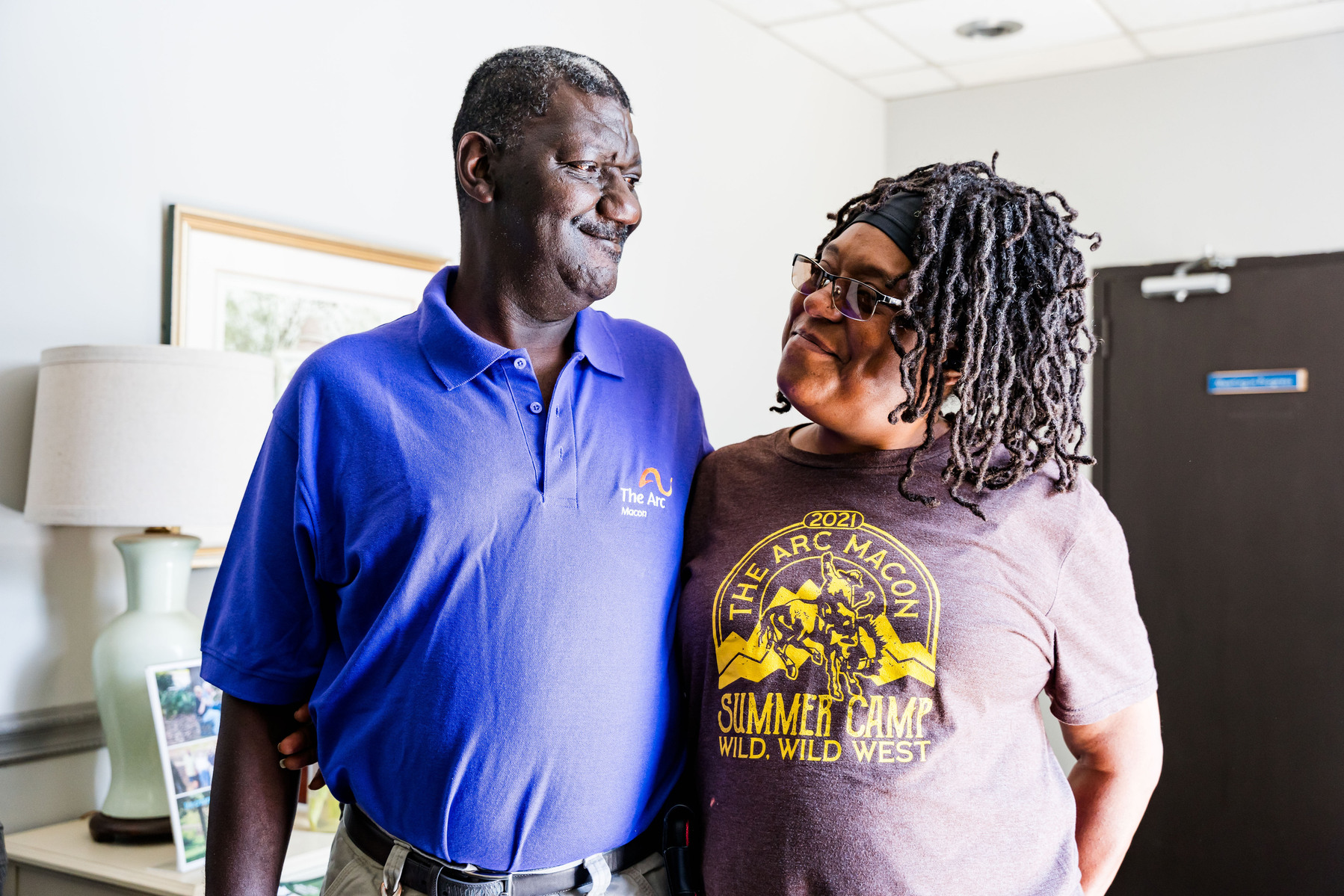 An African American man wearing a purple polo shirt hugs an African American woman with braid and glasses. 