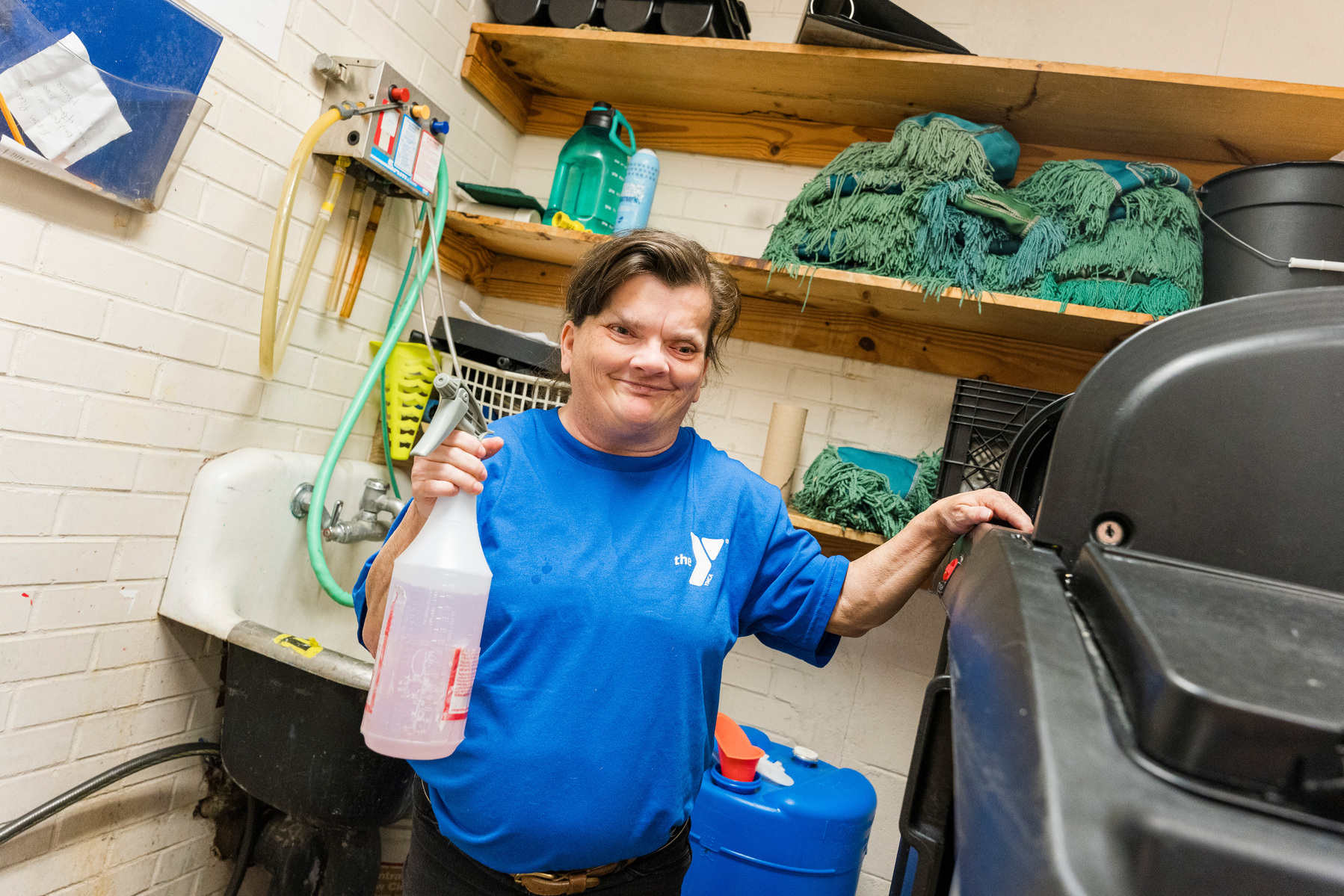 A woman with brown hair wears a blue t-shirt and holds a spray bottle. 