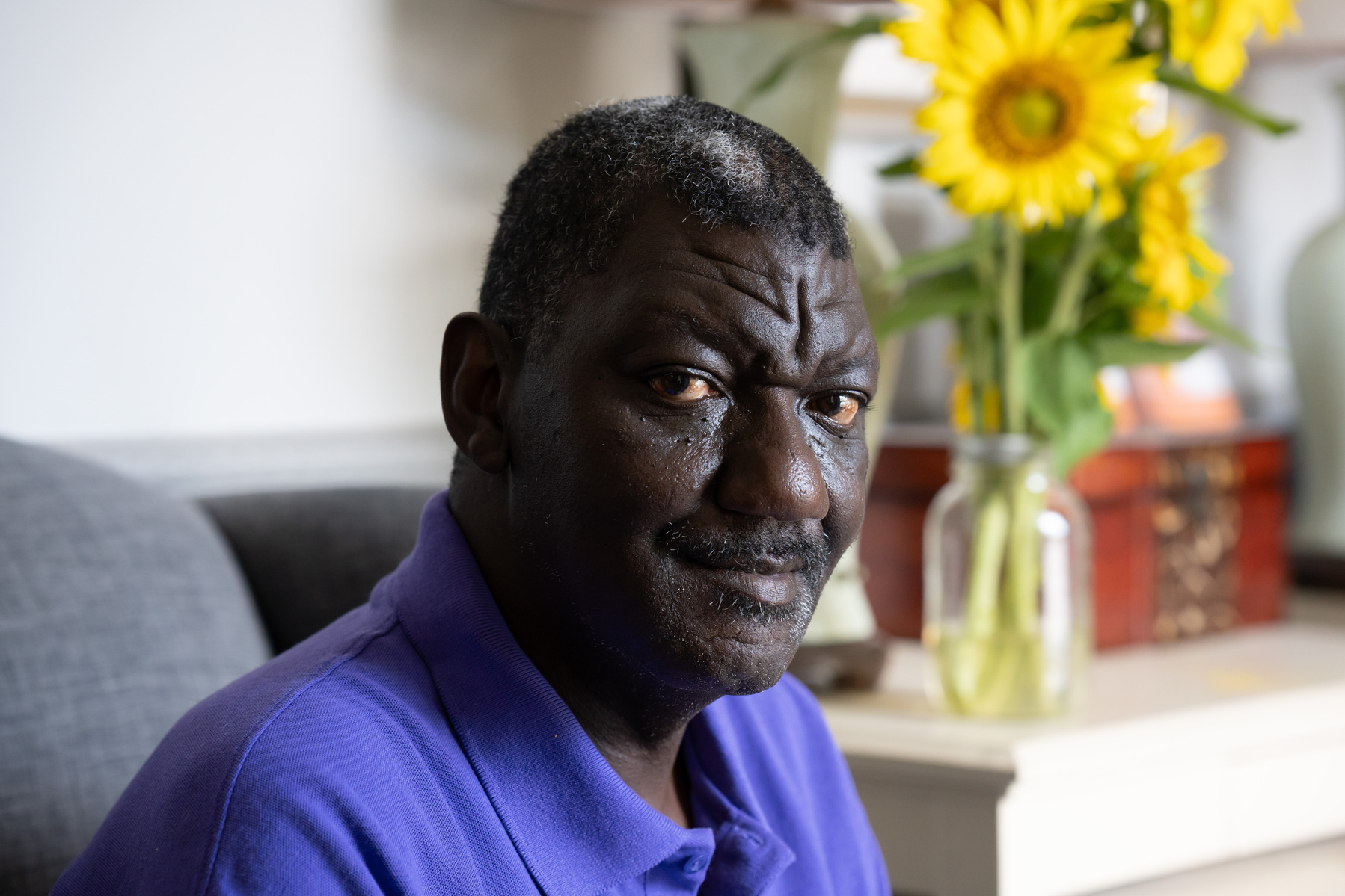 A middle aged African American man wears a purple polo shirt and sits in front of a vase full of sunflowers. 