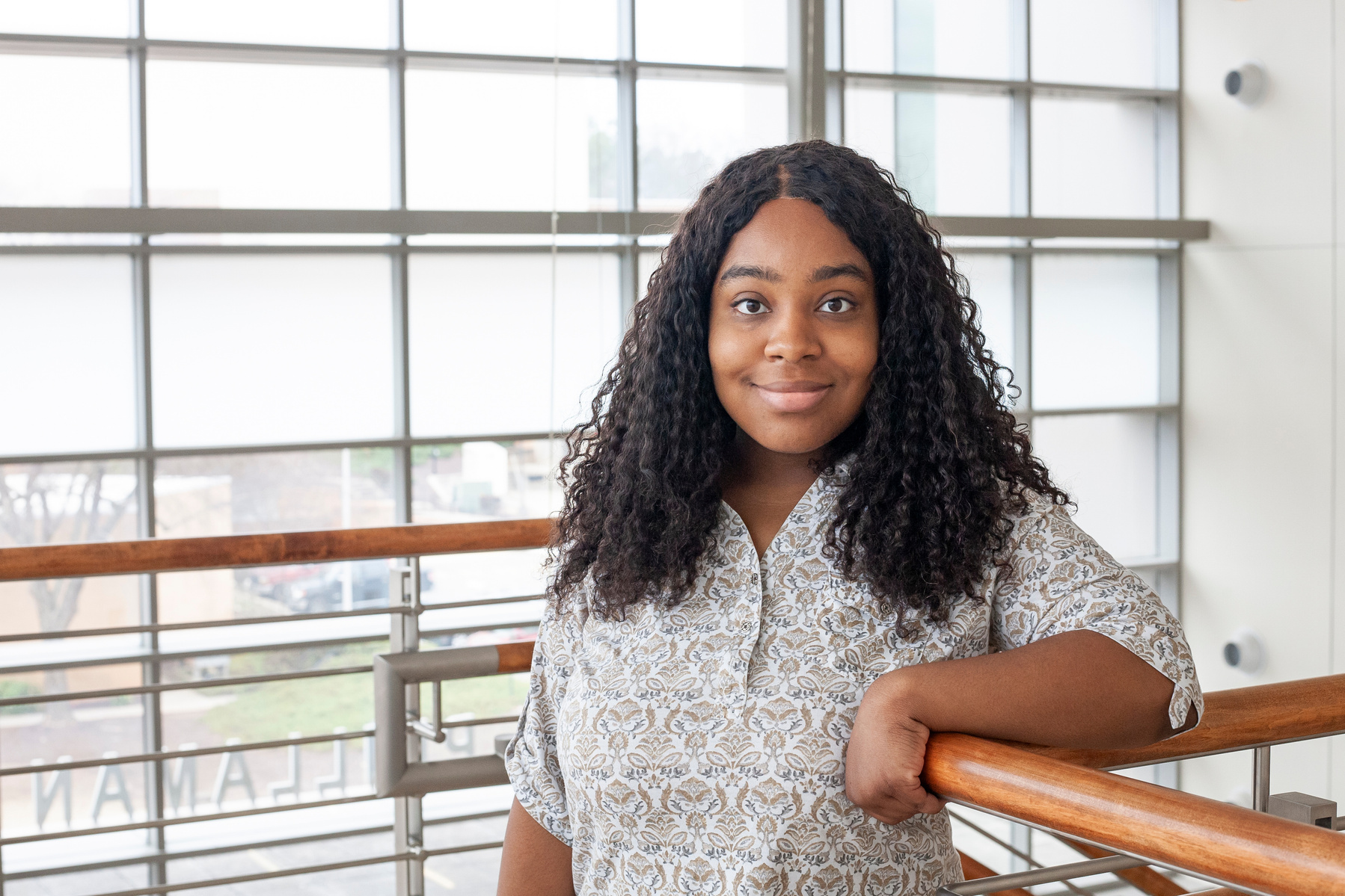 A young African American woman wearing a white blouse leans against a railing in front of a large window. 