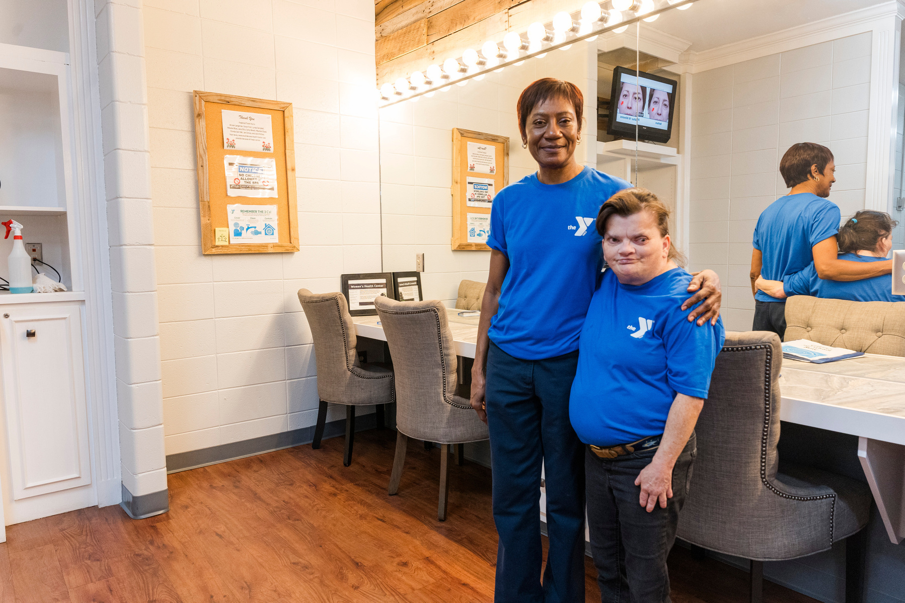 Two women wear matching blue t-shirts and stand in front of a theatre dressing room. 