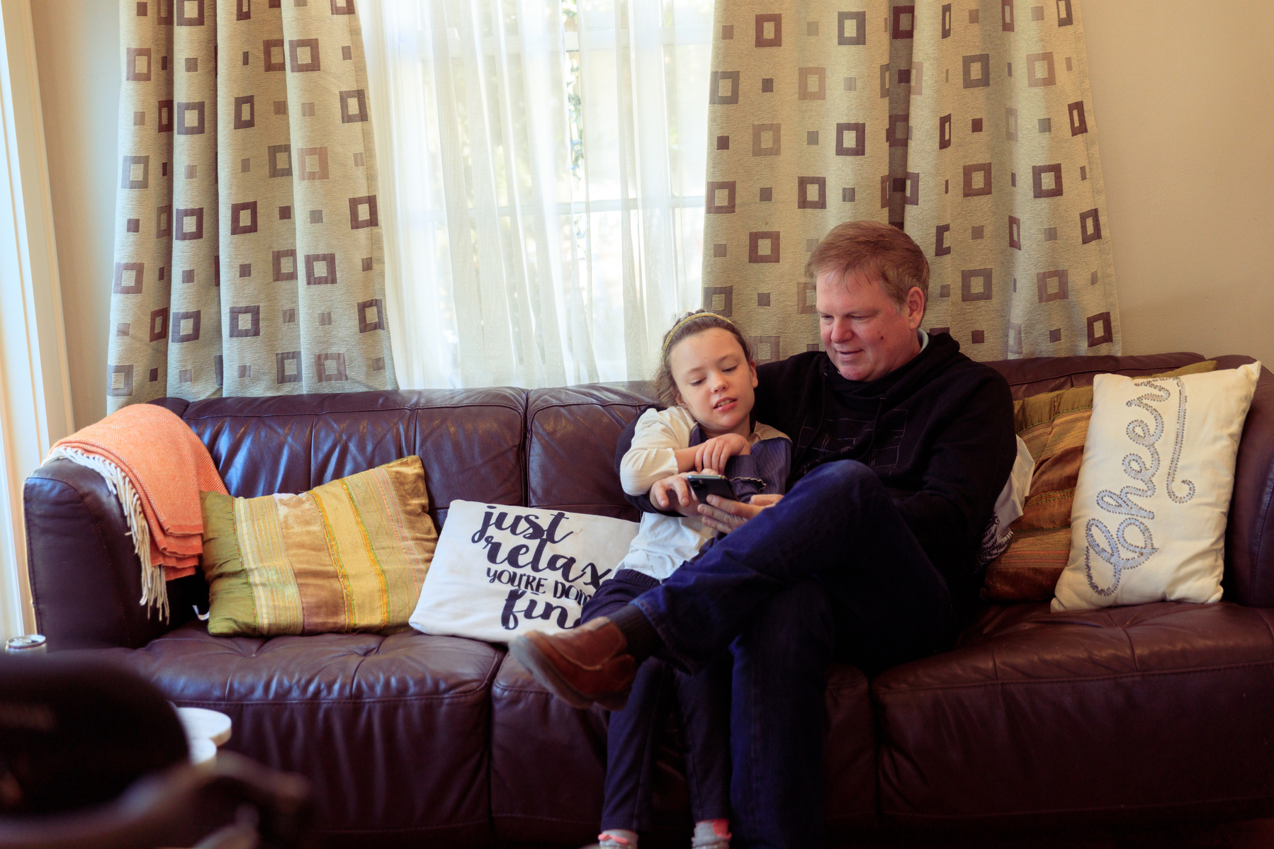 A young girl and her father sit on a sofa and look at a smartphone. 