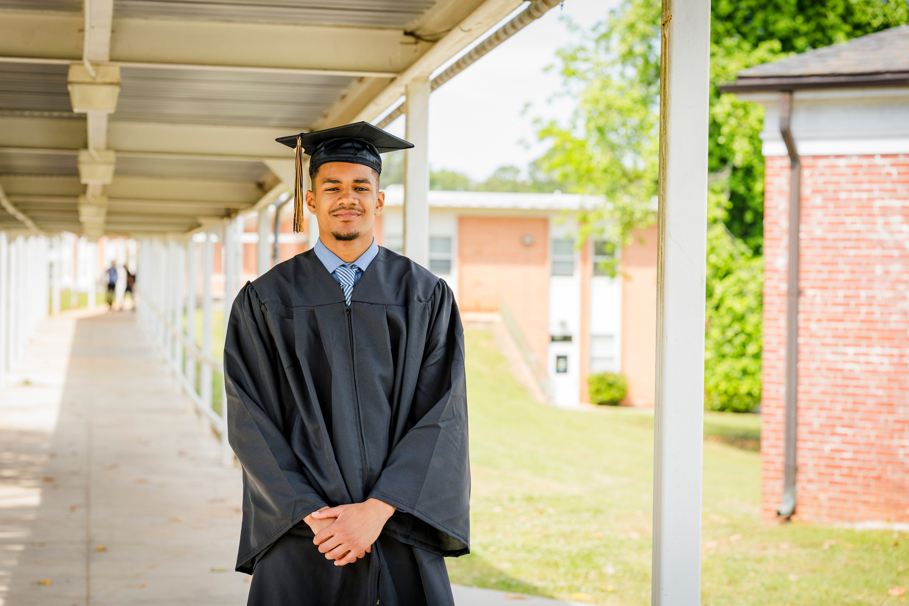 A young African American male wears a graduation cap and gown while standing in a walkway. 