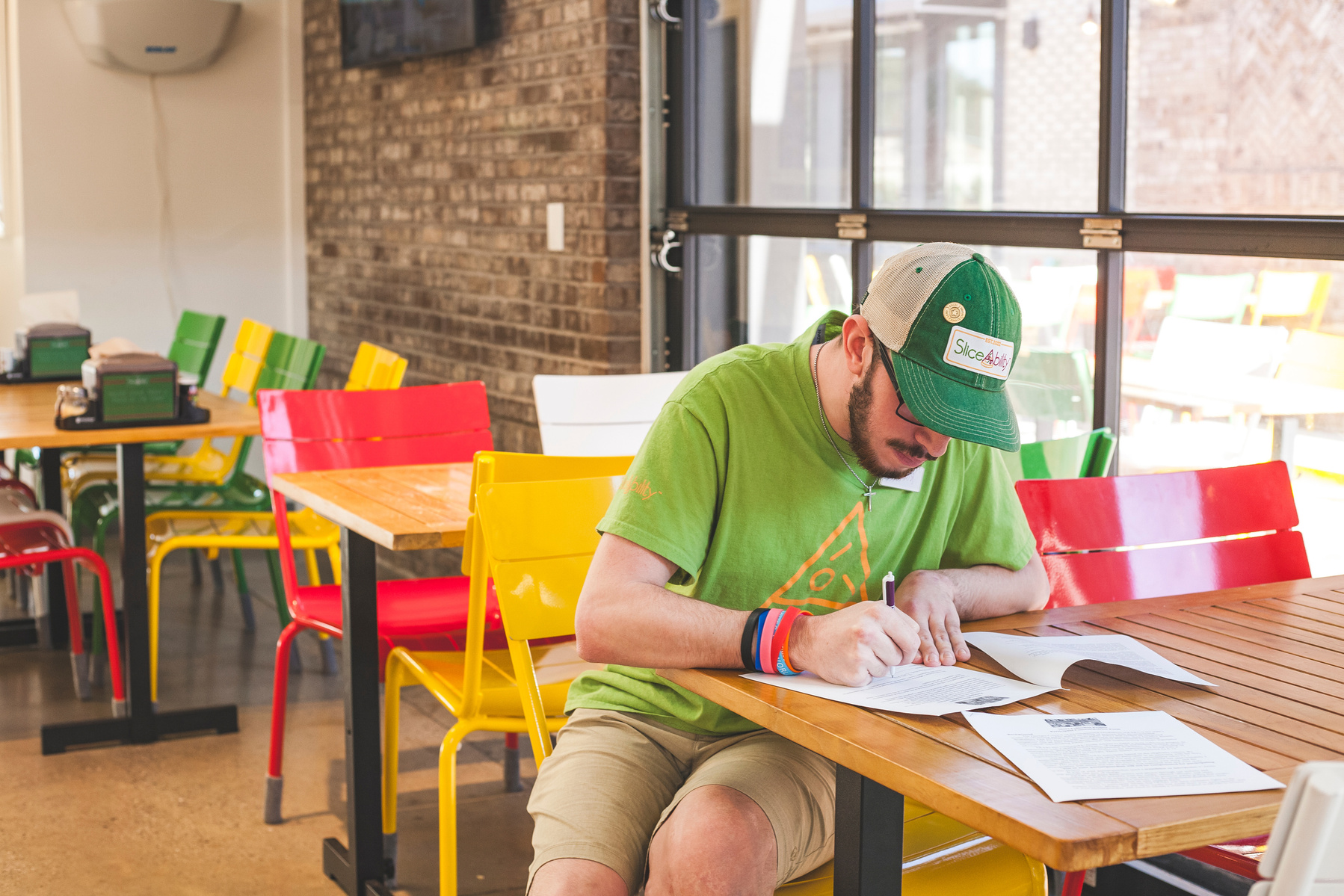A bearded young man wears a green hat and t-shirt works at a restaurant table. 