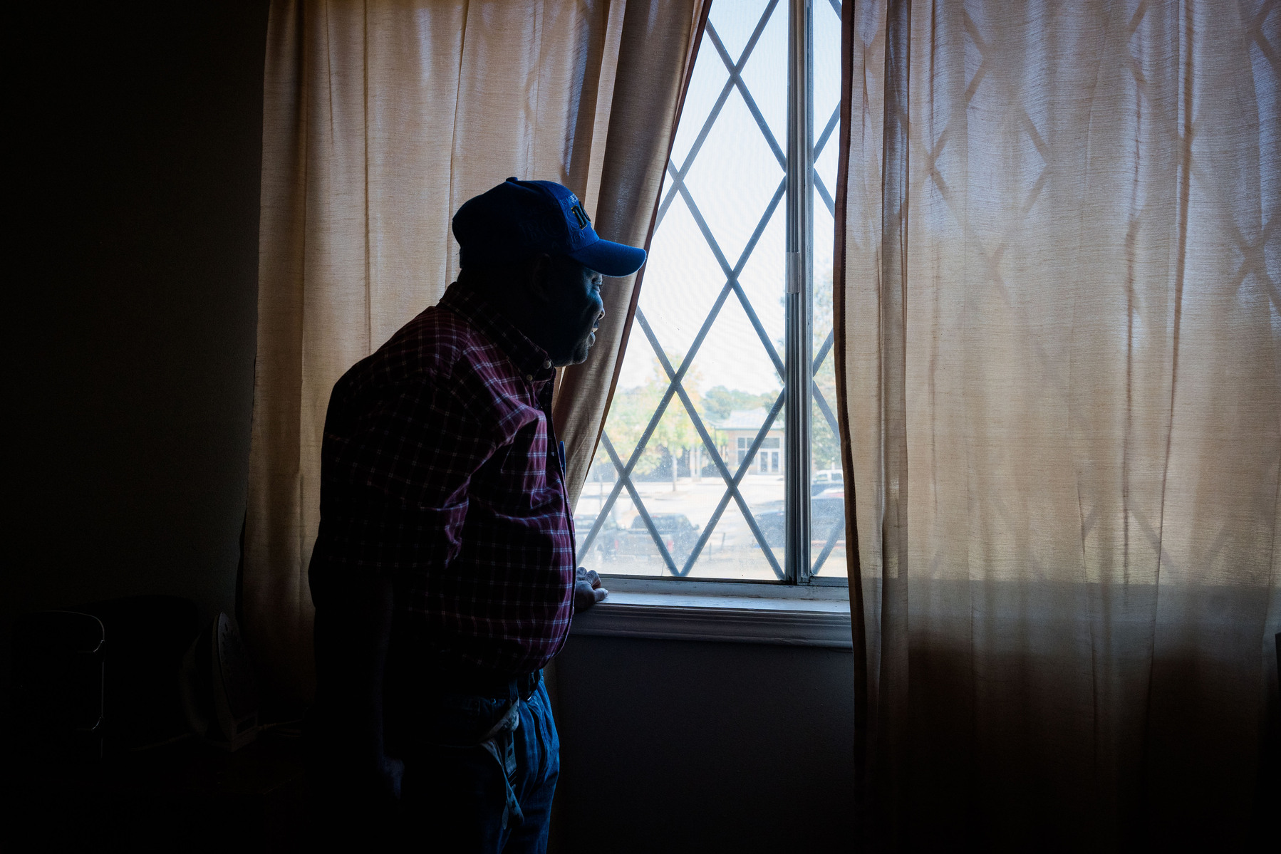 A man wears a plaid shirt and ball cap as he looks out of a window.