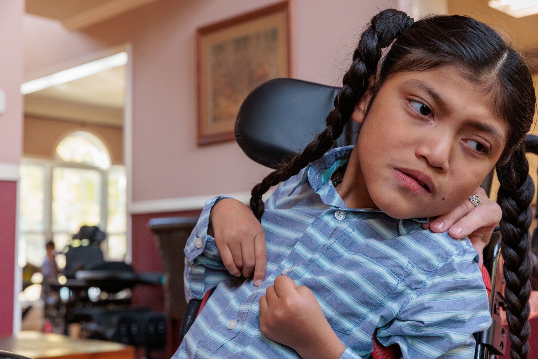 A boy in a striped shirt sits in a wheelchair with his hands in front of him.