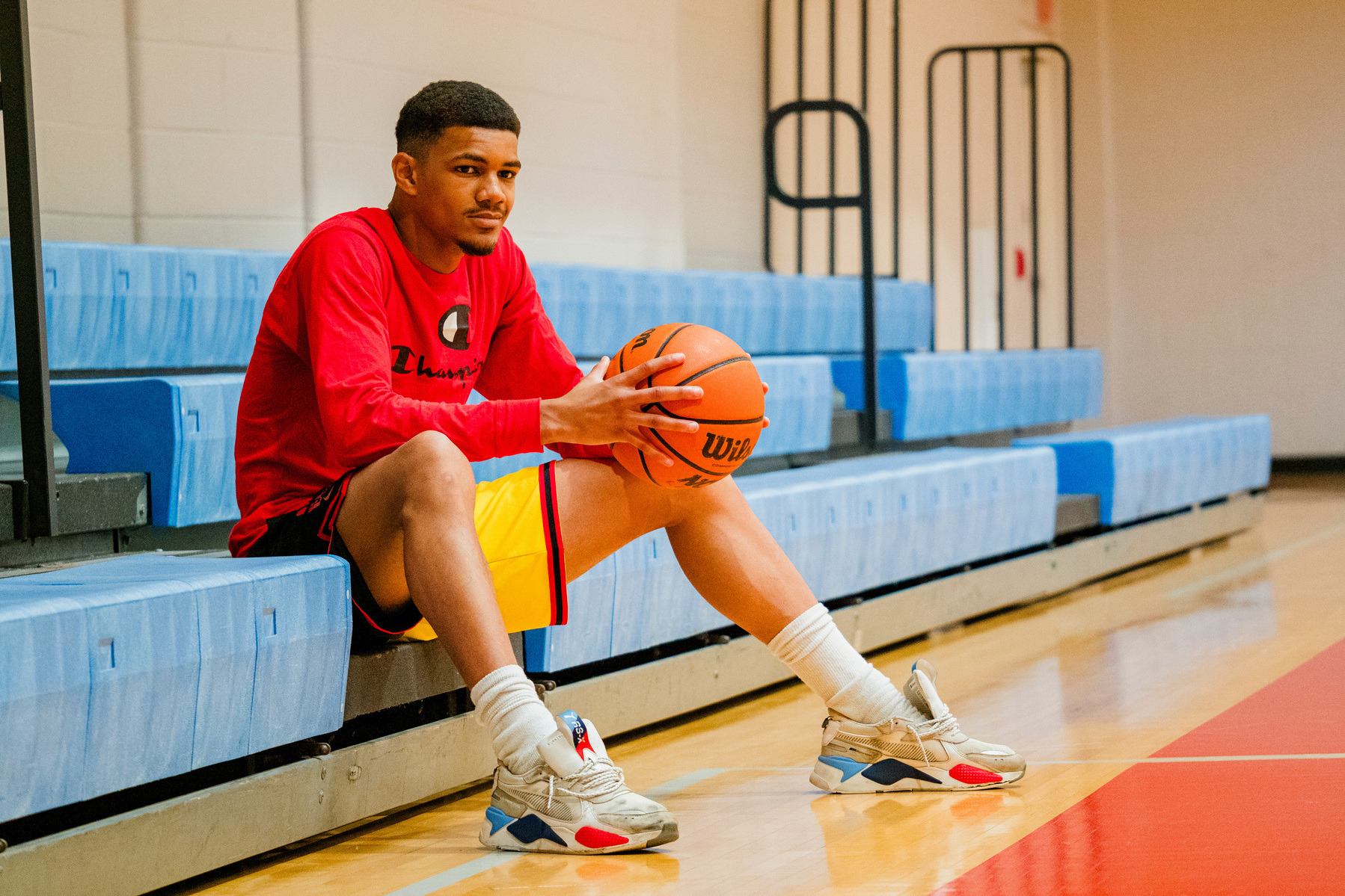 A young African American man sits on bleachers in a gym holding a basketball. 