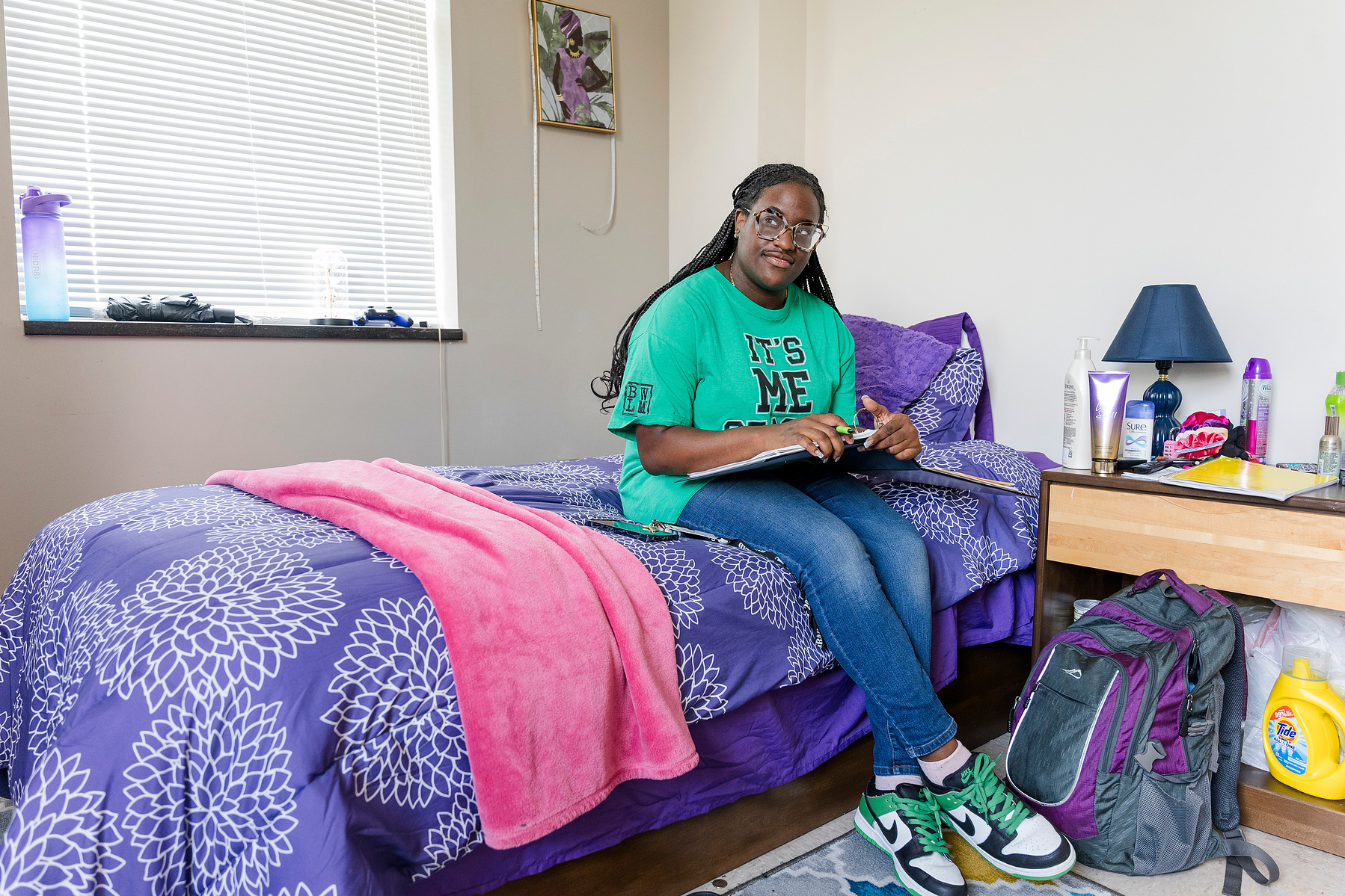 Young African American woman wears braids and a green shirt as she sits on a colorful bed in her dorm room. 