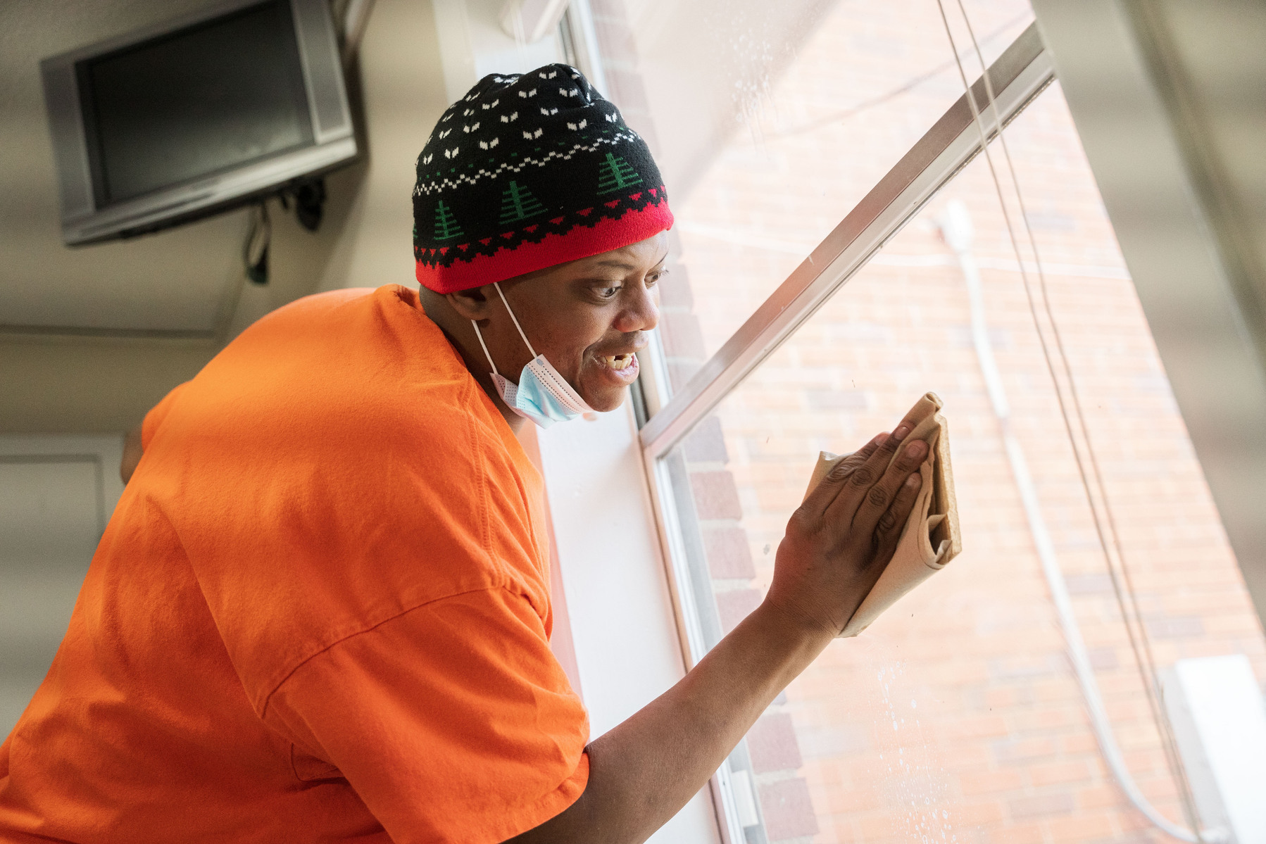 A man in an orange shirt, blue knit cap and white facemask cleans a window. 
